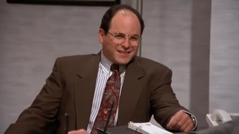 Jason Alexander grins behind his desk as George Costanza in Seinfeld
