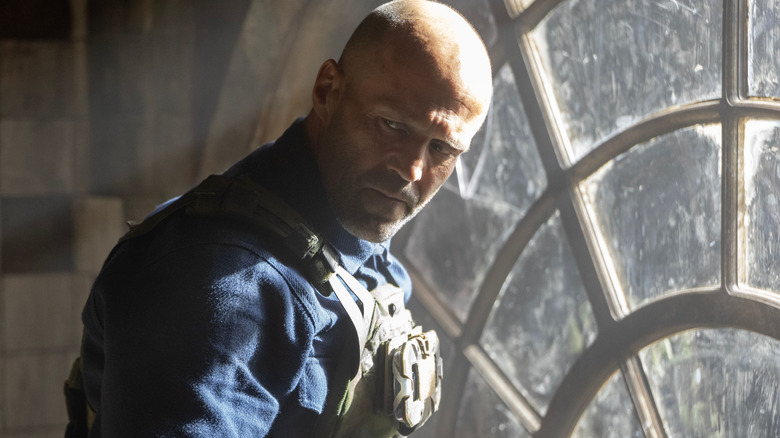 Jason Statham as Levon Cade wearing a tactical vest and standing by a window in A Working Man