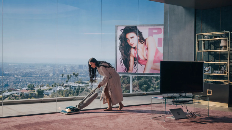 Elisabeth vacuuming in her apartment while a billboard of Sue looks on in The Substance
