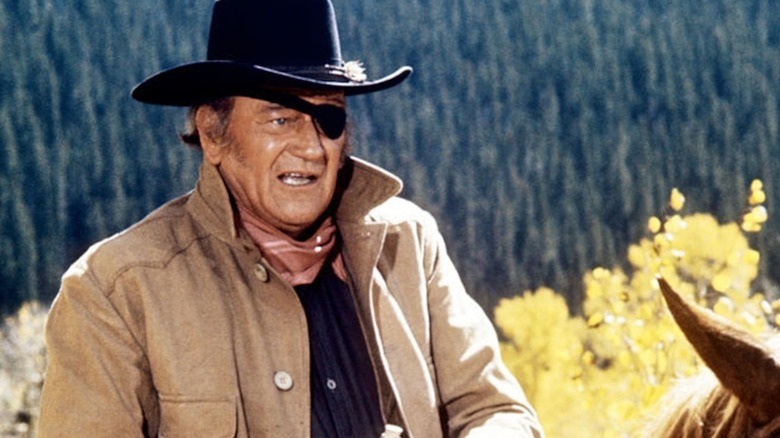 Rooster Cogburn (John Wayne) wears a tan jacket, black cowboy hat, and eye patch while sitting on horseback in front of a forest in True Grit