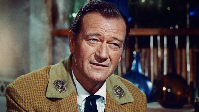 John Wayne's Davy Crockett stands with his arms folded in a shadowy room in The Alamo