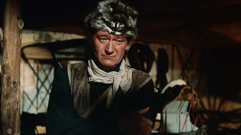John Wayne's Davy Crockett stands with his arms folded in a shadowy room in The Alamo