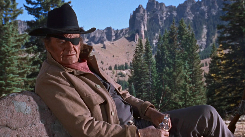 John Wayne as U.S. Marshal Reuben J. "Rooster" Cogburn drinking a bottle of liquor while sitting and leaning on a rock in Rooster Cogburn.