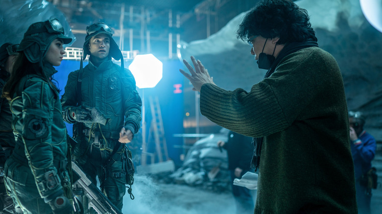 Bong Joon-Ho dirigindo os atores Robert Pattinson como Mickey Barnes e Anamaria Vartolomei como Kai Katz em um bastidores ainda do Mickey 17