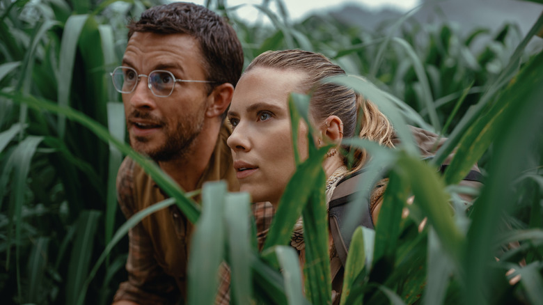 Jonathan Bailey as Henry Loomis and Scarlett Johansson as Zora Bennett crouched together in a field of long grass in Jurassic World Rebirth