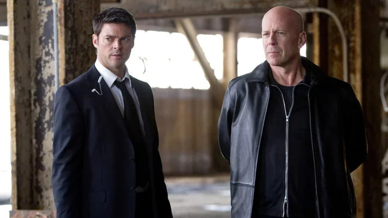 Karl Urban as William Cooper in a black suit and tie and Bruce Willis as Frank Moses in a leather jacket standing in a rusted out building in the movie Red