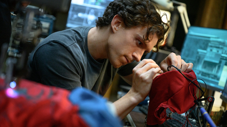Tom Holland as Peter Parker tinkering with his superhero suit at a desk with computer screens in the background in Spider-Man: Brand New Day