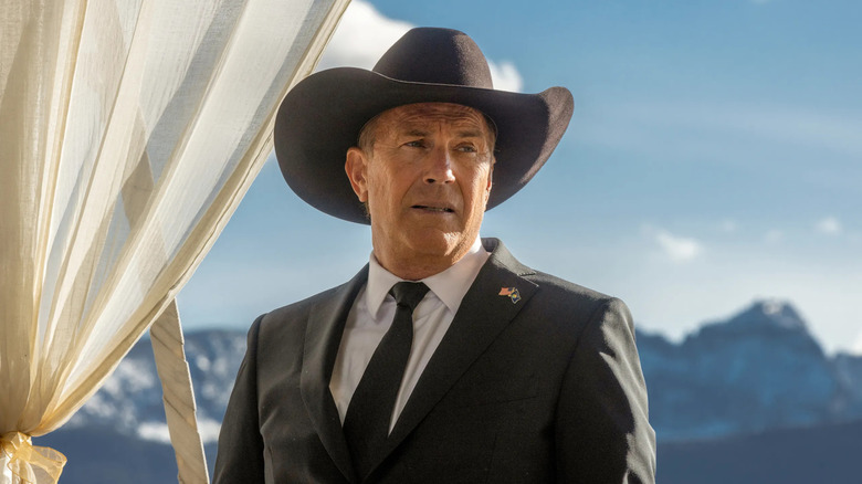 John Dutton (Kevin Costner) looks serious and concerned and wears a black suit and cowboy hat with mountains in the background in Yellowstone