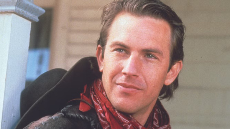 Jake (Kevin Costner) se inclina contra um poste de varanda com um sorriso descontraído, vestindo uma bandana vermelha e chapéu de cowboy preto, em uma cena de Silverado