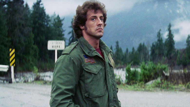 Sylvester Stallone's John Rambo stands on a street in a rural area in First Blood