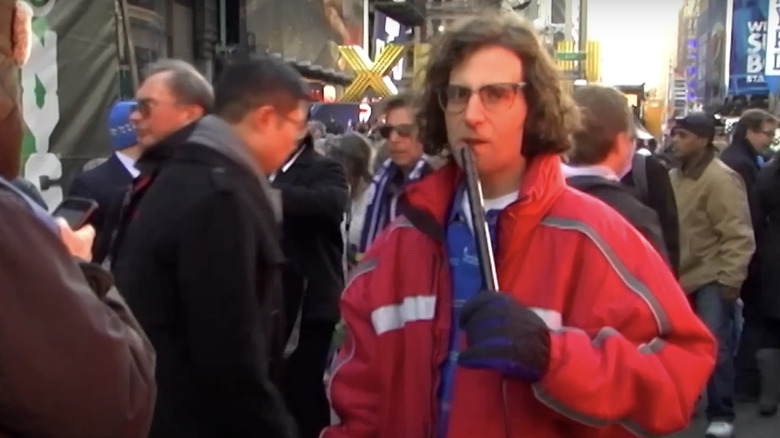 Kyle Mooney speaks into a microphone as he stands, wearing a red coat, filming a report for Saturday Night LIve.