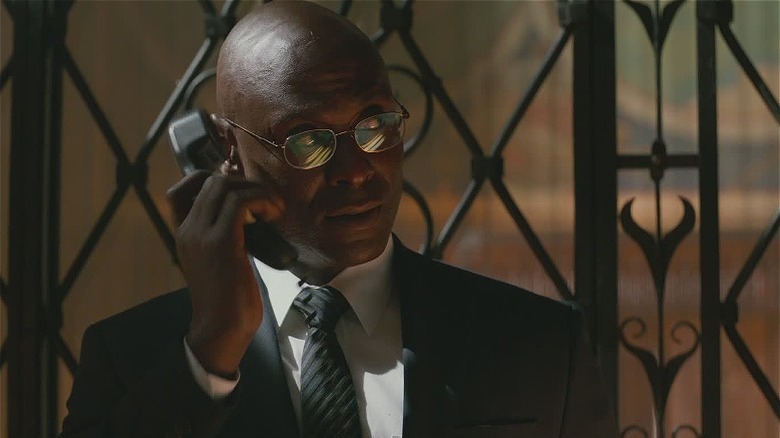 John Wick's Charon holding phone to ear in suit and glasses