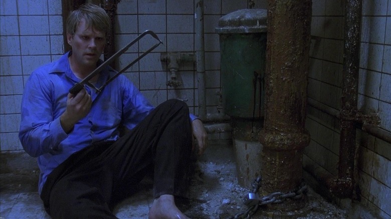 a man looks at a hand saw while sitting on the tile floor next to rusty pipes