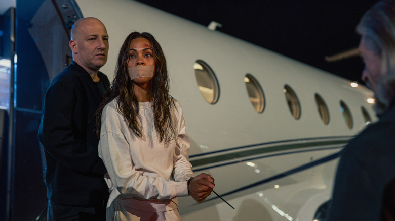 Dimitry Rozental's Renat stands behind Zoe Saldaña's Joe McNamara outside a plane in Lioness