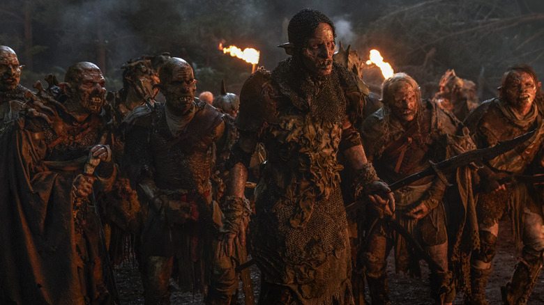 Robert Strange as Glûg holding a spear and standing in formation next to several other orcs dressed in rags in The Lord of the Rings: The Rings of Power