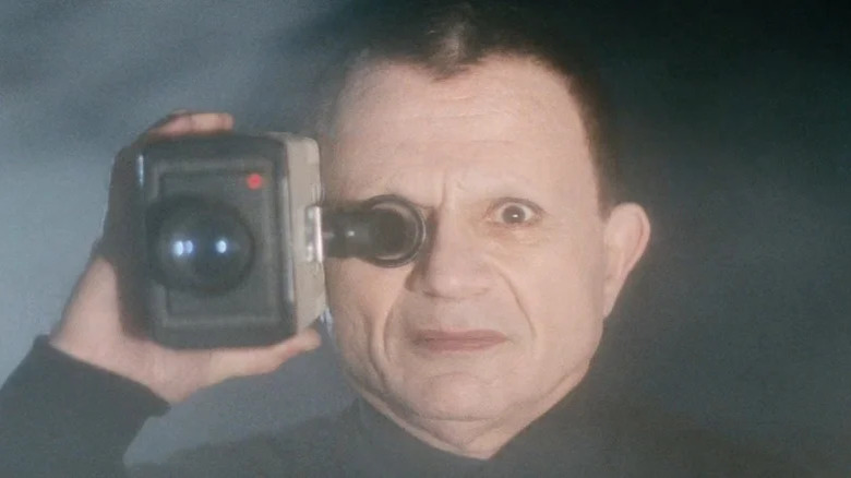 The Mystery Man, holding a camera to his eye in an outdoor, dusty place, a shot from Lost Highway.