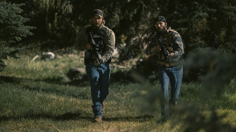 Luke Grimes' Kayce Dutton walks through woodland holding a rifle alongside Logan Marshall-Green's Pete Calvin in Marshals.jpg