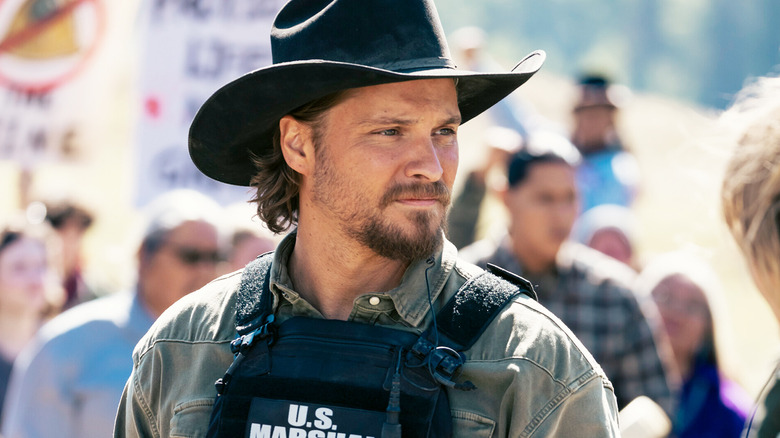 Luke Grimes' Kayce Dutton stands at a protest in a US Marshals vest in Marshals