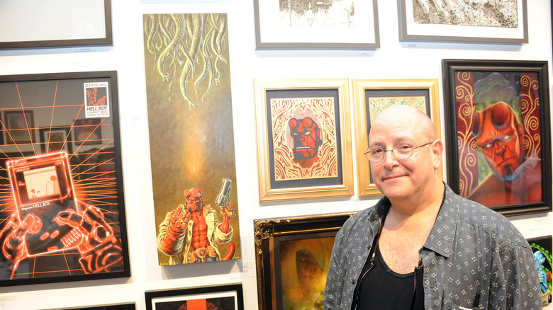 మైక్ మిగ్నోలా ఫ్రేమ్డ్ హెల్‌బాయ్ ఆర్ట్ పక్కన నిలబడి ఉన్నాడు