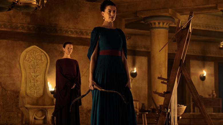 Anne Hathaway as Penelope holding a bow in a firelit room in front of Mia Goth as her maidservant Melantho in The Odyssey