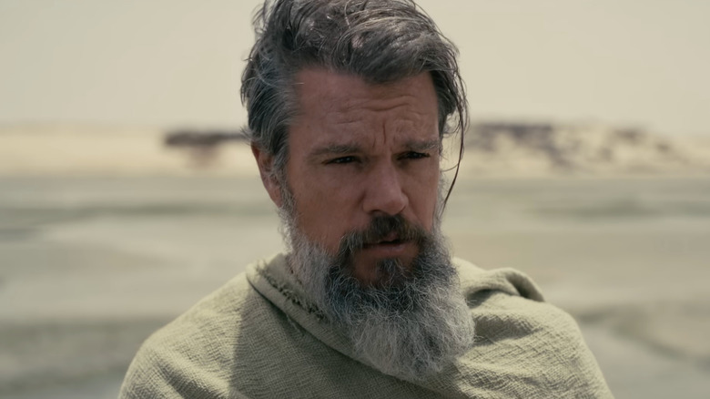 Matt Damon as Odysseus wearing a cloak in front of a sandy area in The Odyssey