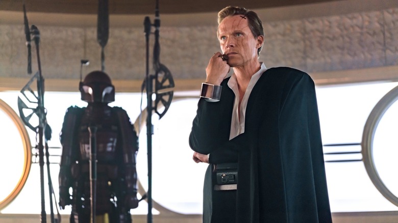Paul Bettany as Dryden Vos wearing a cape and standing next to a suit of Mandalorian armor in Solo: A Star Wars Story