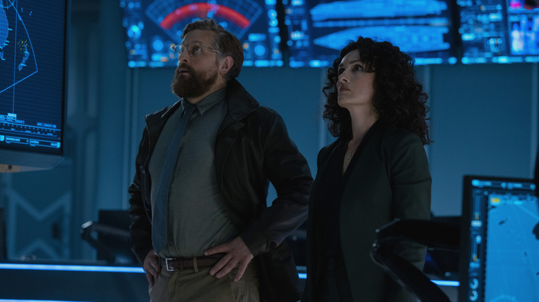 Joe Tippett as Tim standing with his hands on his hips next to Mirelly Taylor as Natalia Verdugo in a room full of screens in Monarch: Legacy of Monsters
