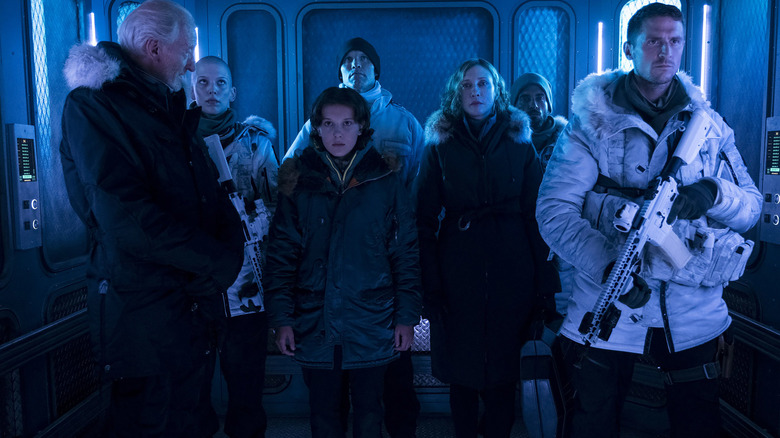 Charles Dance as Alan Jonah standing in an elevator next to Millie Bobby Brown as Madison Russell and Vera Farmiga as Dr. Emma Russell surrounded by armed soldiers in Godzilla: King of the Monsters