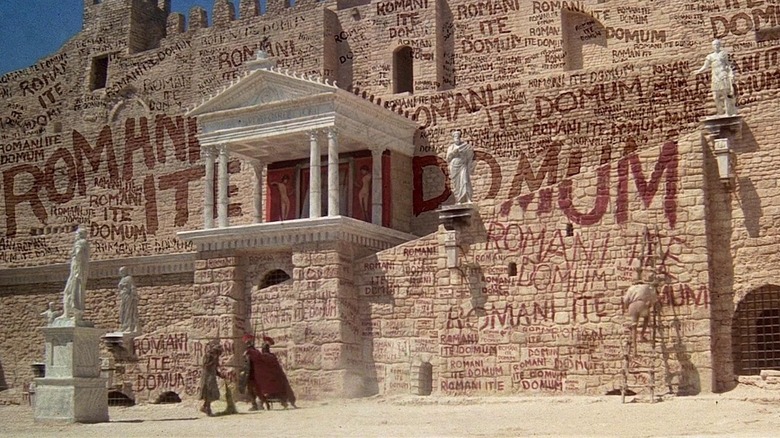 A Roman building covered in graffiti saying 'Romani Ito Domum' in Life of Brian