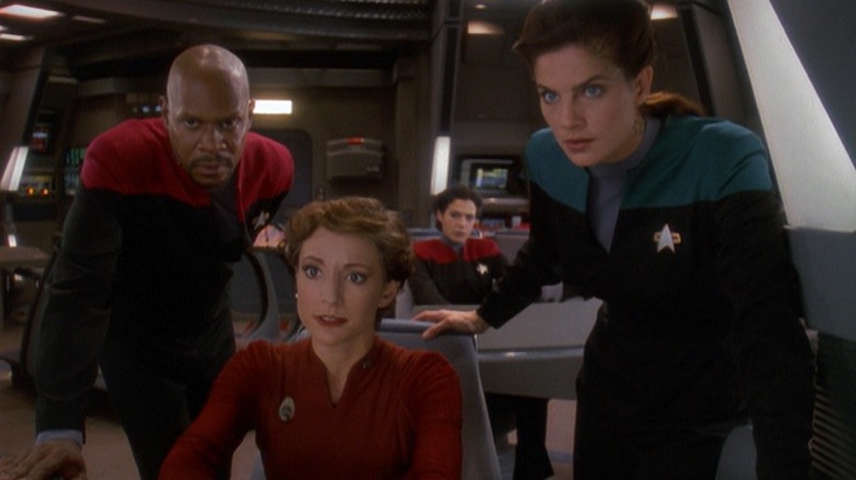 Kira seated at a computer station on the U.S.S. Defiant, flanked by Captain Sisko and Lieutenant Dax