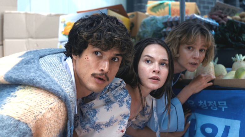 Charles Melton as Austin Davis hiding behind a fruit stand next to Cailee Spaeny as Ashley Miller and Carey Mulligan as Lindsay Crane-Martin in Beef season 2