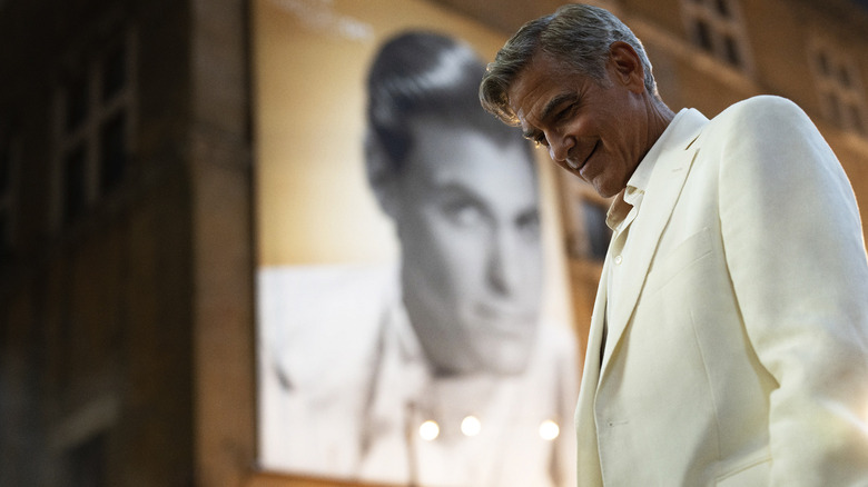 A white suited Jay Kelly smiles while looking down in front of a poster of himself