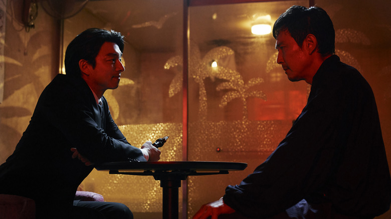 The Recruiter, holding a gun, sits at a table with Seong Gi-hun in Squid Game