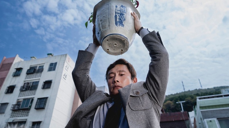 Lee Byung-hun as Man-soo lifting a heavy flower pot over his head from a rooftop in No Other Choice