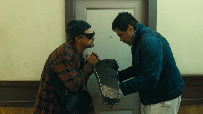 Leonardo DiCaprio as Bob taking a case from Benico Del Toro's Sergio in One Battle After Another