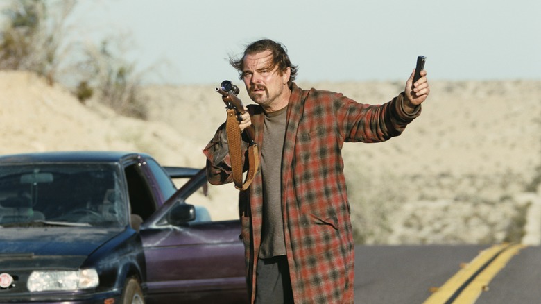 Bob Ferguson walking down a desert road with a scoped rifle in One Battle After Another