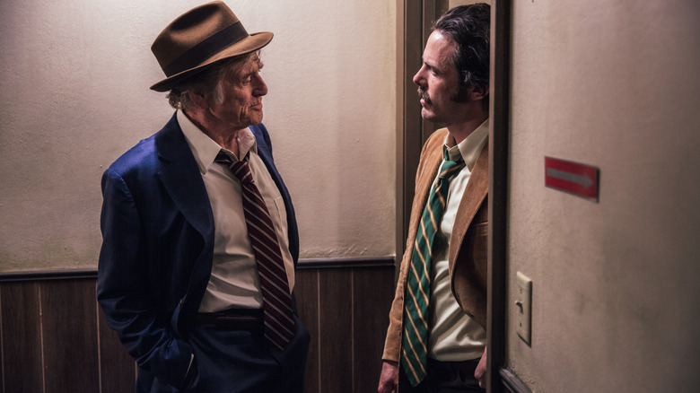 Robert Redford as Forrest Tucker dressed in a top hat and suit standing near a doorway next to Casey Affleck as Detective John Hunt in The Old Man & the Gun
