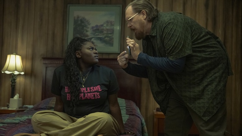 Danielle Brooks as Leota Adebayo sitting on a bed while Steve Agee as John Economos shows her his phone on Peacemaker