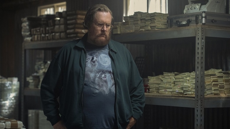 Steve Agee as John Economos looks serious in front of a lot of money, wearing a glorious three raccoon moon shirt