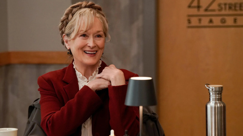 Loretta wearing a red jacket sitting at a table in Only Murders in the Building