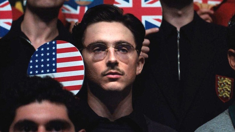 An unsmiling Timotheé Chalamet holds a U.S. flag table tennis paddle as Marty Mauser in Marty Supreme