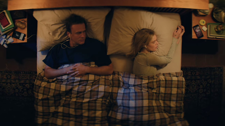 Dan (Jason Segel) and Lisa (Samara Weaving) listening to podcasts while laying in bed in Over Your Dead Body