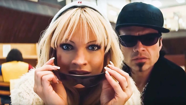 a blonde woman with bangs pulls down sunglasses with two hands to look at something and a man in black with snuglasses and a black cap is behind her on the right