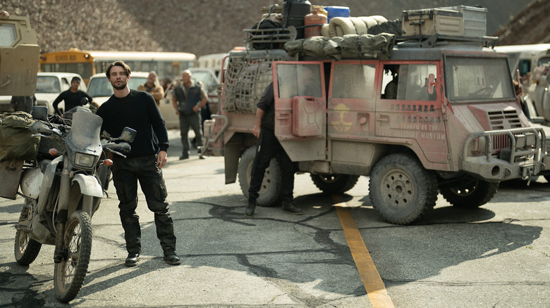 Thomas Doherty as Link standing among a group of followers and dusty vehicles in Paradise season 2