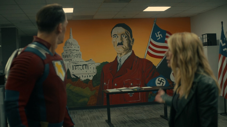 John Cena as Peacemaker and Jennifer Holland as Emilia Harcourt standing in front of a mural of Hitler in front of an American flag with a swastika