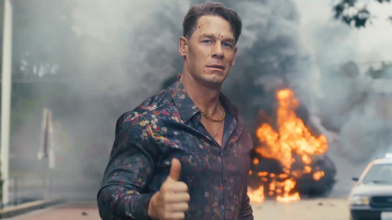 John Cena as Christopher Smith in season 2 of Peacemaker, giving a thumbs up to the camera with a flaming car behind him