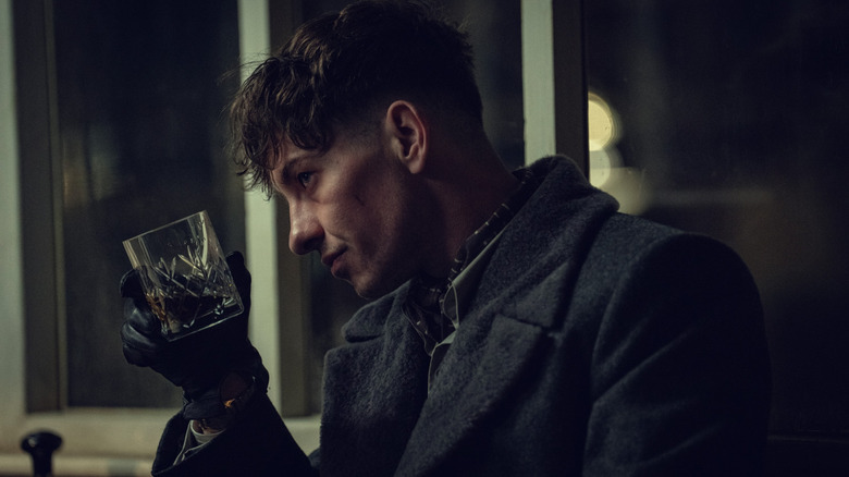 Barry Keoghan als Duke die een jas draagt ​​en een glas whisky omhoog houdt in Peaky Blinders: The Immortal Man