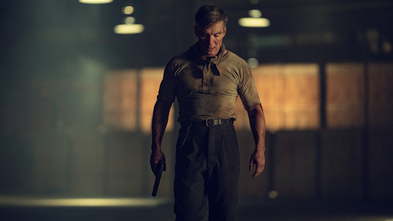 సిలియన్ మర్ఫీ టామీ షెల్బీగా ధూళితో కప్పబడి, చేతిలో తుపాకీ పట్టుకుని పీకీ బ్లైండర్స్: ది ఇమ్మోర్టల్ మ్యాన్‌లోని ఖాళీ గిడ్డంగి గుండా వెళుతున్నాడు