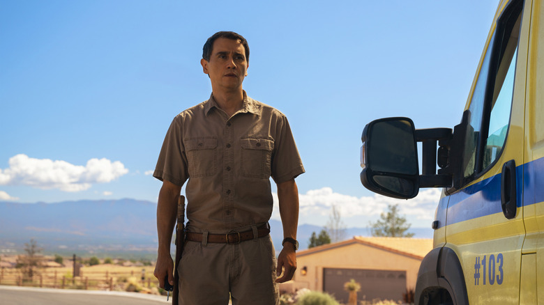 Carlos Manuel Vesga as Manousos standing next to an ambulance on a sunny day in Pluribus