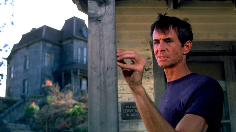 Anthony Perkins in purple shirt in front of motel with looming house in the background in Psycho II